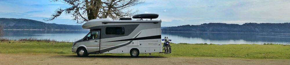 Class C motorhome parked next to a tree with lake, mountain range, and blue sky in the background.