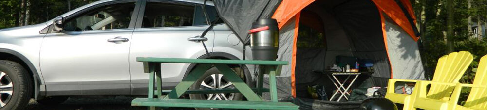 Campsite with tent attached to silver SUV, green picnic table, and yellow lawnchairs.