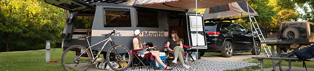 Two women sitting outside a Taxa Mantis travel trailer at a campsite.